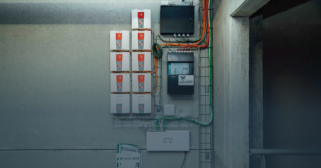 Een uiterst ordelijke technische wandinstallatie op beton door Kristof Devalckenaere, bestaande uit meerdere brandbeveiligingsmodules, een centraal bedieningspaneel en Cisco netwerkapparatuur. De bekabeling is vakkundig weggewerkt in metalen kabeltracés voor een professionele industriële afwerking.