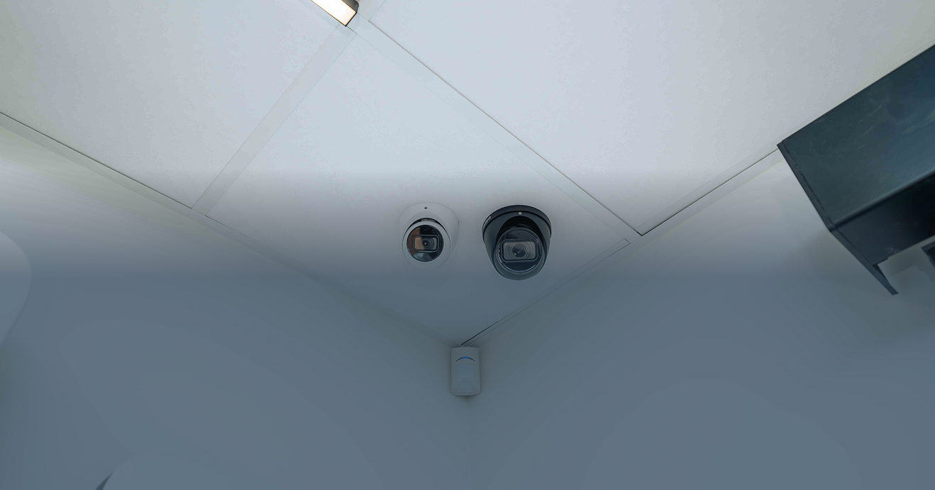 Een blauwe panoramische banner die de integratie van een witte en zwarte dome camera met een bewegingsmelder in een systeemplafond toont, vakkundig geïnstalleerd door Kristof Devalckenaere.