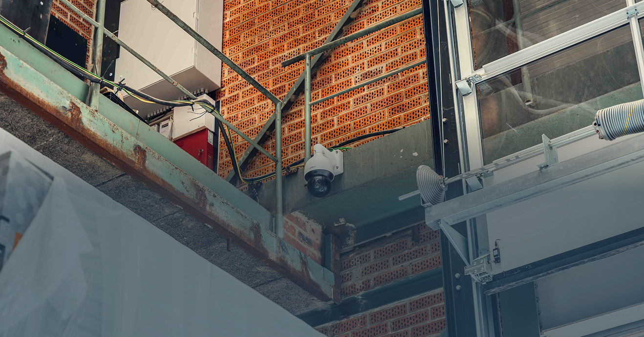 Een witte Dahua dome camera gemonteerd op een stalen constructiebalk in een industriële omgeving met bakstenen muren voor professionele magazijnbeveiliging, vakkundig geïnstalleerd door Kristof Devalckenaere.