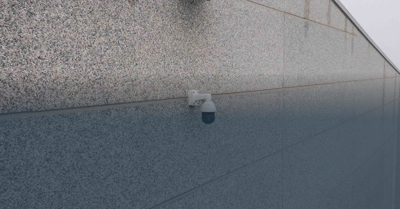 Een close-up foto van een witte professionele dome beveiligingscamera discreet gemonteerd op een moderne, gestructureerde gevel van een gebouw, vakkundig geïnstalleerd door specialist Kristof Devalckenaere voor betrouwbare buitenbeveiliging.