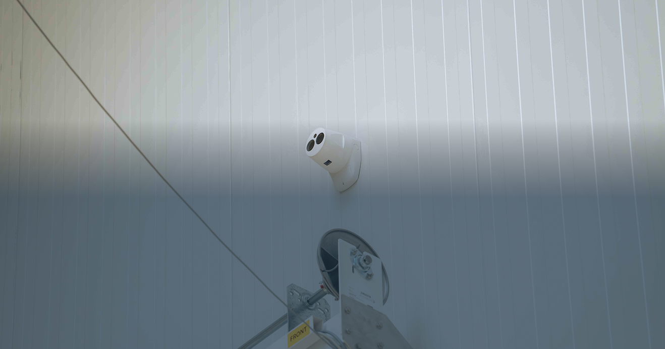 Een panoramische banner met een blauwe filter die een witte multi-sensor camera toont, gemonteerd op een industriële wand voor uitgebreide perimeterbeveiliging, geïnstalleerd door Kristof Devalckenaere.