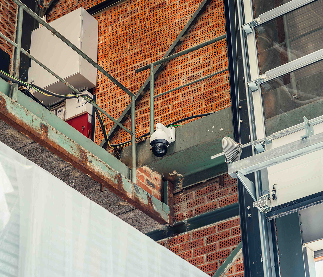 Een witte Dahua dome camera gemonteerd op een stalen constructiebalk in een industriële omgeving met bakstenen muren voor professionele magazijnbeveiliging, vakkundig geïnstalleerd door Kristof Devalckenaere.