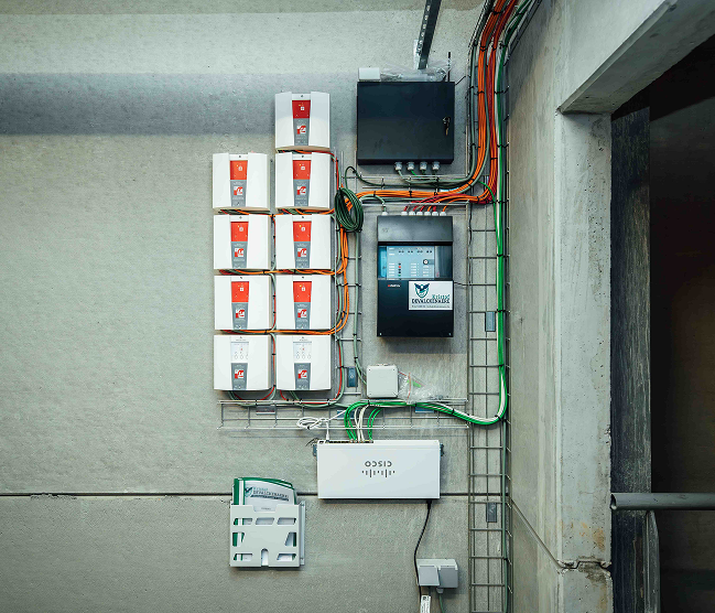 Een uiterst ordelijke technische wandinstallatie op beton door Kristof Devalckenaere, bestaande uit meerdere brandbeveiligingsmodules, een centraal bedieningspaneel en Cisco netwerkapparatuur. De bekabeling is vakkundig weggewerkt in metalen kabeltracés voor een professionele industriële afwerking.