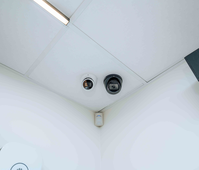 Beveiligingsinstallatie in een modern kantoorgebouw in Oostkamp, uitgevoerd door Kristof Devalckenaere. De foto toont twee CCTV dome-camera's en een PIR-sensor gemonteerd op het systeemplafond in een hoek, met een gegraveerde metalen plaque in het midden die het project identificeert.
