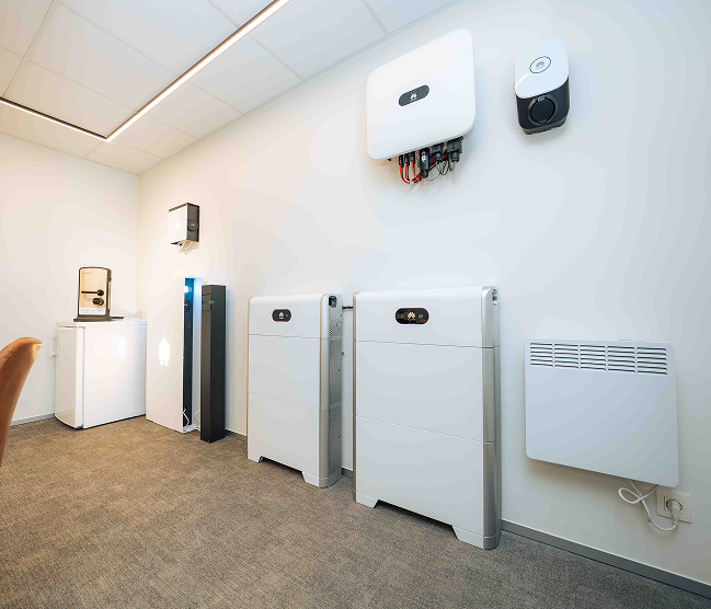 Een weids overzicht van de professionele geïntegreerde systemen demonstratiewand door Kristof Devalckenaere. De foto toont de volledige Huawei LUNA2000 energie-opslag oplossing, inclusief de SUN2000 omvormer en meerdere batterijstacks op de vloer. Daarnaast is een deel van de beveiligingshub zichtbaar met de multi-sensor detector en de toegangsfobs, samen met een wandconvector. Alles strak gemonteerd voor een holistic zicht op de aangeboden building solutions.