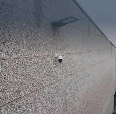 Een close-up foto van een witte professionele dome beveiligingscamera discreet gemonteerd op een moderne, gestructureerde gevel van een gebouw, vakkundig geïnstalleerd door specialist Kristof Devalckenaere voor betrouwbare buitenbeveiliging.