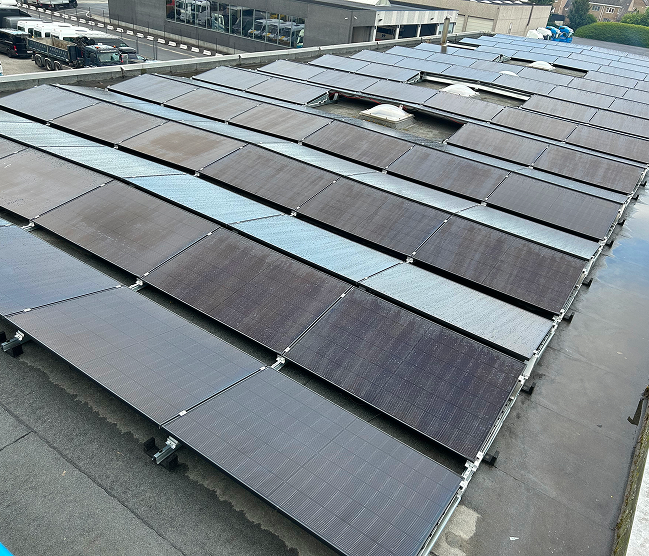 Een overzichtsfoto van een omvangrijke zonnepanelen installatie op het platte dak van een bedrijfspand, met een wagenpark en trucks in de achtergrond. Gerealiseerd door specialist Devalckenaere Kristof.