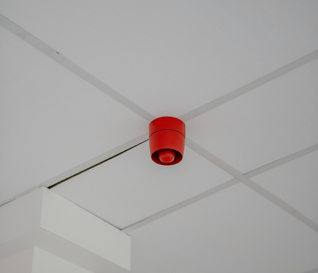 Een felrode brandmeldalarmsirene (sounder) vakkundig gemonteerd op een wit systeemplafond als onderdeel van een professionele branddetectie installatie door Kristof Devalckenaere.