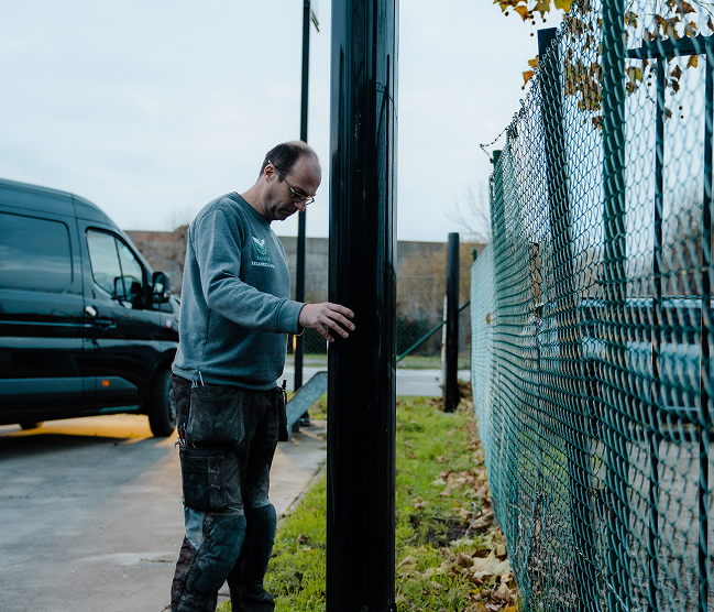 Installateur Kristof Devalckenaere aan het werk bij de installatie van een automatische poortpaal. De technicus in Devalckenaere werkkleding inspecteert de motor of bediening van een geautomatiseerd hekwerk, met een werkbus op de achtergrond. Focus op professionele poortautomatisatie.