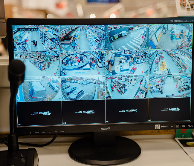 Een monitor toont live beelden van meerdere Dahua beveiligingscamera's in een winkel, inclusief parkeerplaats en gangpaden, geconfigureerd door Kristof Devalckenaere.